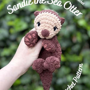 May include: A crocheted sea otter plush toy named Sandie the Sea Otter, featuring a dark brown body, a lighter beige face, and dark brown embroidered eyes. The text Sandie the Sea Otter and Snuggler Crochet Pattern is visible on the image.