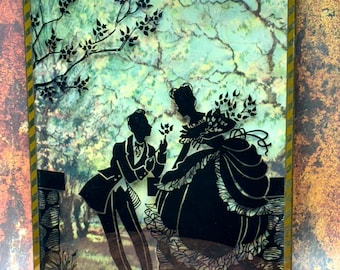 Puede incluir: Obra de arte de silueta enmarcada que representa una escena romántica. Las siluetas negras de un hombre y una mujer se colocan sobre un fondo suave, similar a una acuarela, de color verde y azul. El marco tiene un borde a rayas doradas y blancas.