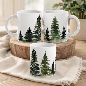Peut inclure: Trois mugs en céramique blanche avec un motif aquarelle de pins verts. Les mugs sont disposés sur une surface en bois et un tissu beige. Les mugs ont une anse standard et une forme cylindrique.