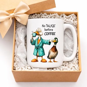 Peut inclure: Tasse en céramique blanche avec un motif de canard humoristique, présentée dans une boîte cadeau avec un nœud. La tasse représente un canard en peignoir buvant du café, avec le texte "No talkie before coffee."
