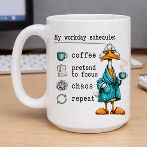May include: White ceramic mug with a humorous illustration of a goose in a robe holding a coffee cup. The mug has the text "My workday schedule: coffee, pretend to focus, chaos, repeat."