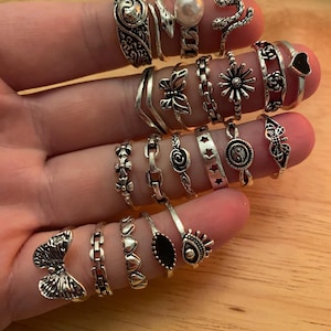 May include: A selection of silver rings, each with a unique design, displayed on fingers. The rings include floral, butterfly, snake, and heart shapes. Some rings feature a pearl or a black stone. The rings are presented on a light brown surface.