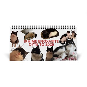 May include: A spiral-bound desk calendar featuring various images of cats in different poses. The calendar has the text "ΜΗ ΜΕ ΕΝΟΧΛΕΙΤΕ ΟΥΤΕ ΤΟ 2026" in red letters. The cats are black and white, and some are wearing jewelry.