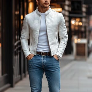 May include: A white textured leather jacket with a stand-up collar and zip closure. The jacket is worn with a white t-shirt, blue jeans, and a brown belt. A silver watch is visible on the wrist.