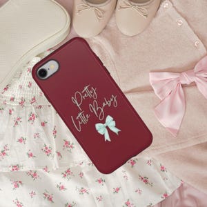 Könnte beinhalten: Burgunderfarbene Handyhülle mit dem Schriftzug "Pretty Little Baby" in Schreibschrift und einer hellblauen Schleife. Die Hülle ist von einem rosa Cardigan, Ballerinas und einem Blumenkleid umgeben.