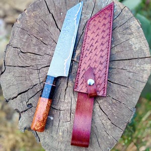 May include: A handmade chef's knife with a patterned steel blade and a wooden handle with black accents. The knife rests on a wooden surface next to a burgundy leather sheath with a woven pattern.