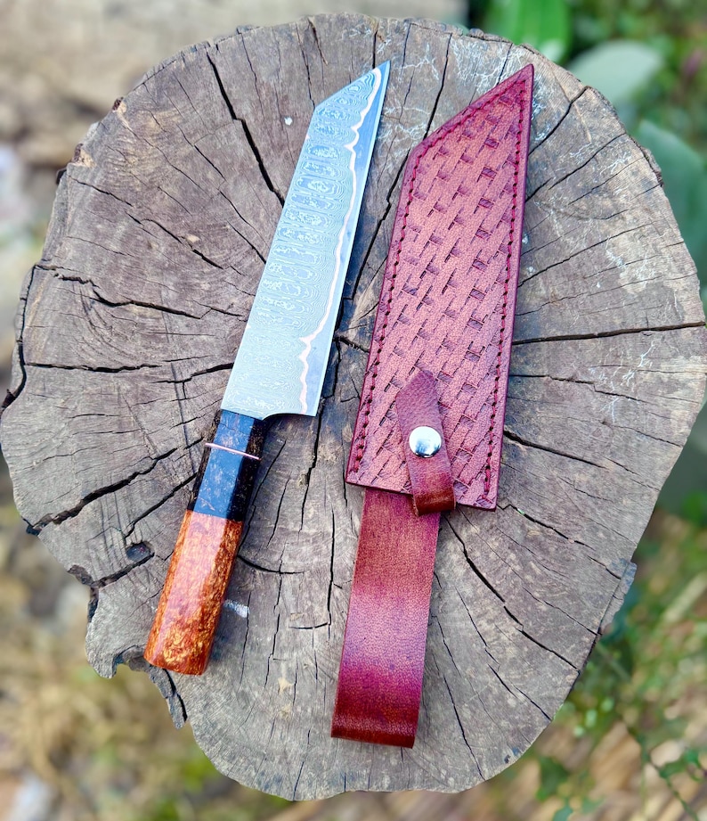 May include: A handmade chef's knife with a patterned steel blade and a wooden handle with black accents. The knife rests on a wooden surface next to a burgundy leather sheath with a woven pattern.