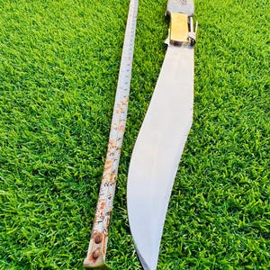May include: A large, curved blade knife with a decorative handle and brass accents. A metal measuring tape is placed next to the knife. The knife blade is approximately 12 inches long.