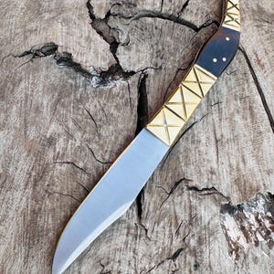 May include: A handcrafted knife with a curved, silver blade. The handle features a dark wood grip, accented with gold-colored metal and decorative geometric patterns. The knife rests on a weathered wooden surface.