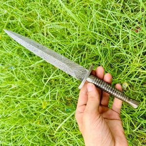 Puede incluir: Una daga de acero de Damasco con una hoja estampada y un mango acanalado. La daga se sostiene sobre un fondo de hierba verde.