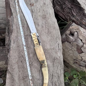 Puede incluir: Un cuchillo grande y ornamentado con una hoja curva y un mango de hueso. El mango está adornado con bandas de metal dorado. Una cinta métrica se coloca junto al cuchillo. El cuchillo mide unos 40 cm de largo.