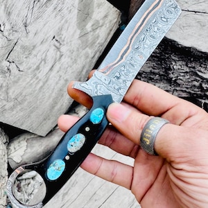 Puede incluir: Una navaja recta con una hoja de acero estampada y un mango negro con incrustaciones de piedras turquesas. La hoja tiene detalles en cobre. La navaja se sostiene sobre un fondo de madera envejecida.