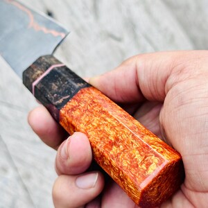 May include: Close-up of a handcrafted knife with a blade and a handle. The handle is a vibrant orange and red, with a unique wood grain pattern. The blade has a silver finish with a copper-colored accent.