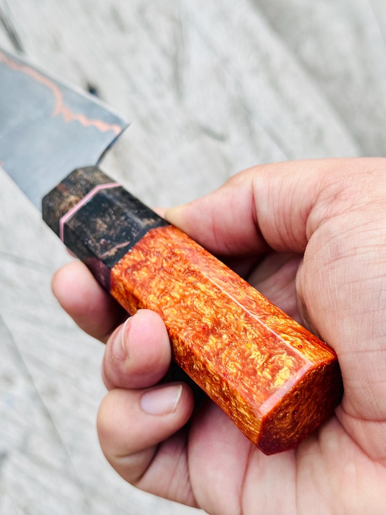 May include: Close-up of a handcrafted knife with a blade and a handle. The handle is a vibrant orange and red, with a unique wood grain pattern. The blade has a silver finish with a copper-colored accent.