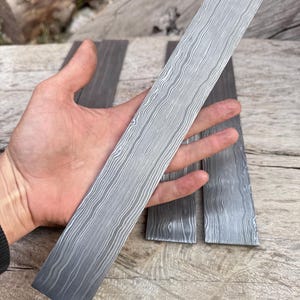 May include: A long, rectangular piece of steel with a unique, wavy pattern. The steel has a gradient of gray tones, from dark at the bottom to lighter at the top. Two additional pieces of steel are visible in the background, also with the same pattern.