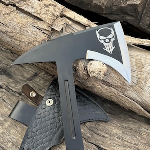May include: A black tactical tomahawk featuring a silver skull graphic on the blade and a sharp, pointed spike on the opposite side. The tool includes a black leather sheath with a textured scale pattern and metal snap button closures.