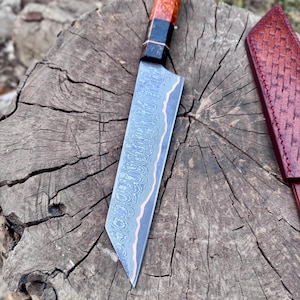 May include: A Damascus steel chef's knife with a patterned blade and a wooden handle with red and black accents. The knife rests on a weathered wooden surface, with a leather sheath visible in the background.