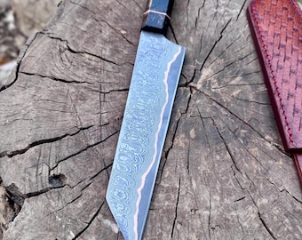 May include: A Damascus steel chef's knife with a patterned blade and a wooden handle with red and black accents. The knife rests on a weathered wooden surface, with a leather sheath visible in the background.