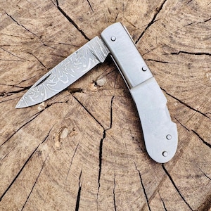 May include: A partially open, silver folding knife with a patterned blade. The knife is set against a textured, brown wooden background, showcasing the blade's intricate design and the knife's overall metallic finish.