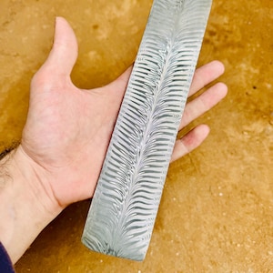 Puede incluir: Una pieza rectangular de acero con un diseño único. El patrón es una serie de líneas curvas, creando una apariencia de pluma. El acero tiene una combinación de colores plateado y gris. La pieza se sostiene en una mano.