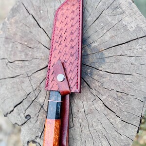 May include: A handcrafted knife with a patterned, reddish-brown leather sheath. The knife handle features a unique wood grain design with black and orange accents. The sheath is secured with a metal snap.