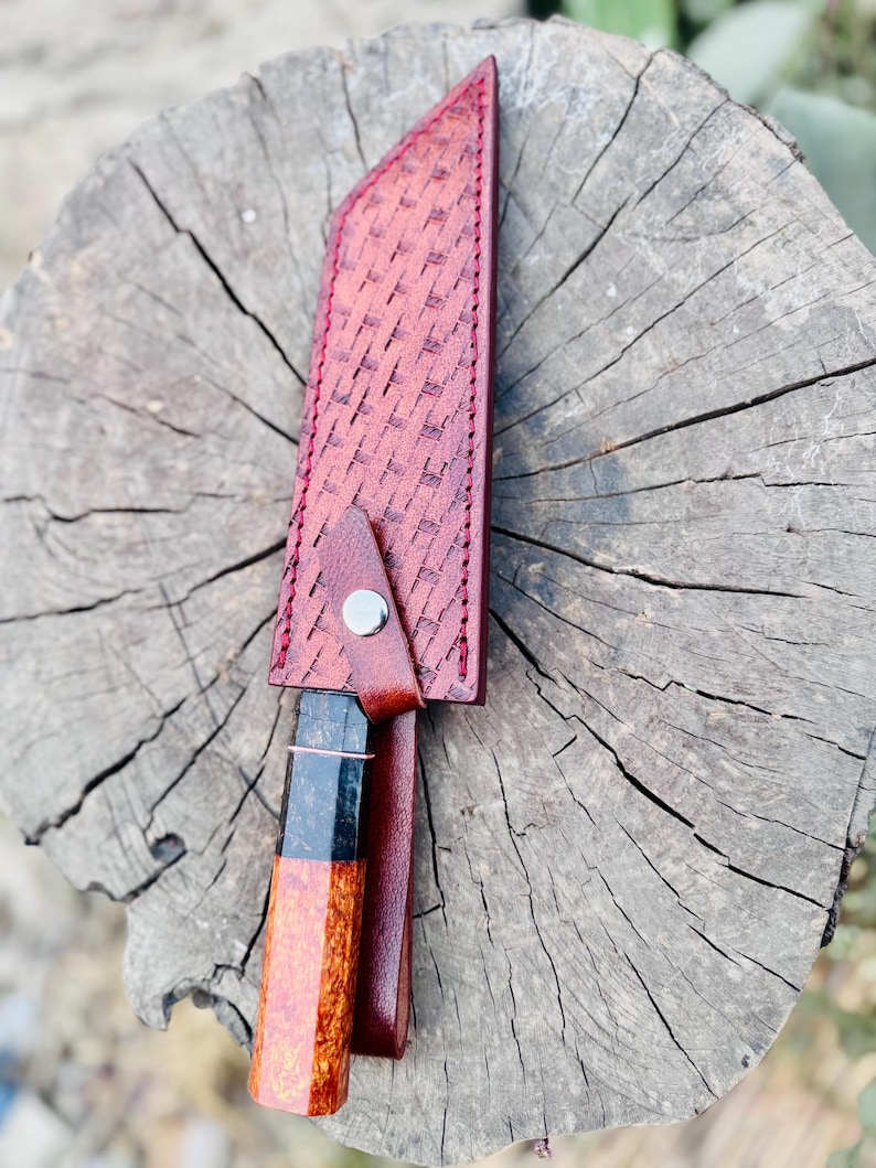 May include: A handcrafted knife with a patterned, reddish-brown leather sheath. The knife handle features a unique wood grain design with black and orange accents. The sheath is secured with a metal snap.