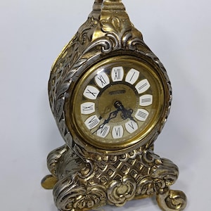May include: An antique gold and silver-toned clock with a decorative design. The clock face has Roman numerals and the word "SPLENDOR" printed on it. The clock has a decorative base and a pointed top.