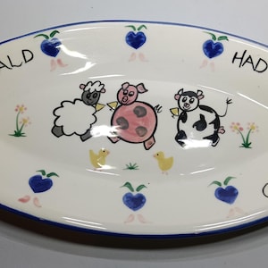 Może przedstawiać: Owalny ceramiczny półmisek z niebieską obwódką i czarnym napisem "Old McDonald Had a Farm". Półmisek przedstawia kreskówkowe ilustracje owcy, świni i krowy, a także dwa żółte pisklęta i niebieskie jagody w kształcie serca.