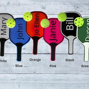 Pode incluir: Uma coleção de seis raquetes de pickleball coloridas com bolas amarelas a condizer. Cada raquete é de uma cor diferente: branco, azul, laranja, rosa, preto e verde. Cada raquete tem um nome impresso: Mary, John, Jo Ellen, Janie, Bill e Duncan.