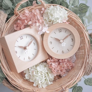 以下が含まれることがあります： ピンクと白の花が入った柳のバスケットの中に、明るい文字盤と花のアクセントが付いた2つの木製時計があります。1つの時計は四角く、もう1つは丸いです。時計には黒い数字と針があります。