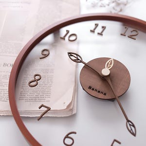 May include: A circular wall clock with a brown wooden frame and a transparent face. The clock has wooden numerals and leaf-shaped hands. The centre of the clock features a wooden circle with the word "Banana".