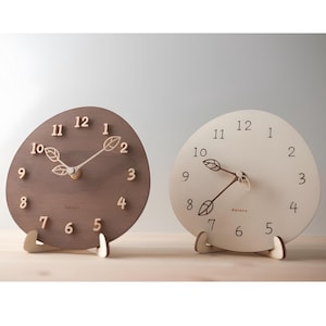 May include: Two wooden analog clocks. One is brown with beige numbers and hands, the other is off-white with brown numbers and hands. Both have a leaf design at the 10 o'clock position and sit on wooden stands.