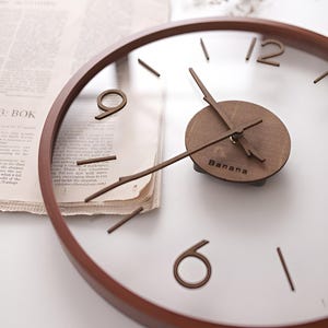 May include: A round wall clock with a clear face and a brown wooden frame. The clock features brown wooden hands and hour markers. The word "Banana" is printed on the clock face.