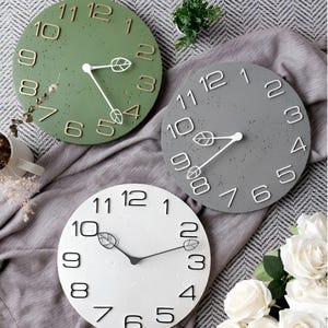 May include: Three circular wall clocks in green, grey, and white. Each clock has a textured finish, leaf-shaped details, and contrasting numerals. The hands are white or black. These clocks are decorative and suitable for home decor.
