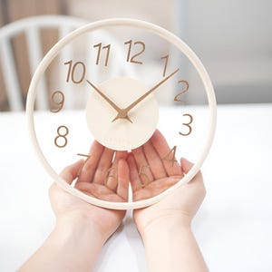 May include: A circular, transparent clock with a light wood frame. The clock face has brown numerals and hands, with a cream-coloured centre. The clock is held by two hands.