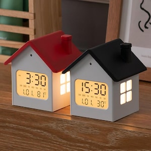 May include: Two house-shaped digital alarm clocks with white bodies and chimneys. One has a red roof, the other a black roof. Both feature illuminated LCD screens displaying time, date, and temperature in Celsius, along with a glowing window design.