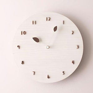 May include: A circular, white wooden clock with brown numerals and leaf-shaped hands. The clock face has a light wood grain texture. The numbers are in a simple, modern font. The clock is designed for wall mounting.