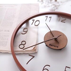 May include: A circular wall clock with a brown wooden frame and hands. The clock face is transparent with brown numerals. The clock's centre features a wooden circle with the word "Banana" printed on it.
