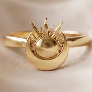 May include: A gold-colored ring featuring a sun and crescent moon design. The ring's band is smooth and rounded, with the sun and moon forming a central, detailed pendant. The sun has radiating rays, and the moon cradles the sun.