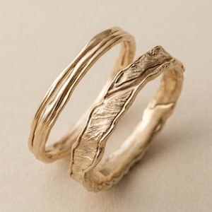 May include: Two gold rings with textured, organic designs. One ring has parallel lines, the other a rough, bark-like texture. The rings are set against a neutral background, highlighting their unique craftsmanship and design.