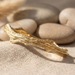 May include: A gold-colored ring with a textured, organic design resembling a tree branch. The ring is set against a neutral background with smooth stones and a textured fabric, creating a natural aesthetic.