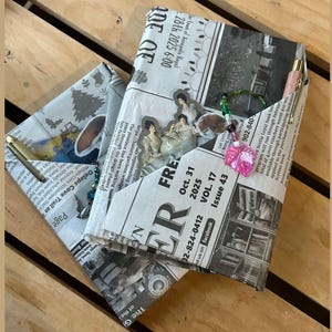 Può includere: Diario fatto a mano ricoperto di giornale, con una penna e perline decorative. Il diario presenta una tasca con il testo "FREE" e "Oct. 31 2025". Il diario è su una superficie di legno.