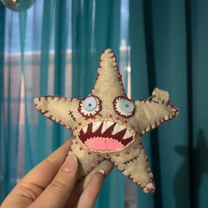 May include: A beige felt star-shaped plush toy with a stitched design. The toy features a wide open mouth with sharp teeth, blue eyes, and red accents. The toy is approximately 10 cm wide.