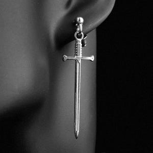 May include: A silver sword-shaped earring is displayed against a black background. The earring is a detailed miniature of a sword, with a crossguard and a pointed blade. The earring is attached to a post and is worn on an ear.