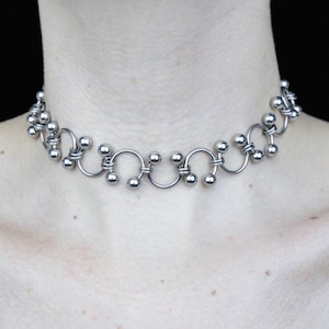 Peut inclure: Un collier ras du cou argenté au design unique. Il présente une série d'éléments en forme de U reliés, chacun orné de petites perles rondes. Le métal a une finition polie, créant une surface réfléchissante. Un bijou élégant.