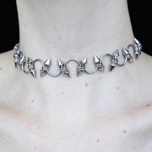 May include: A silver-colored choker necklace featuring a series of pointed spikes. The spikes are evenly spaced and connected by curved metal links. The necklace is designed to sit around the neck.