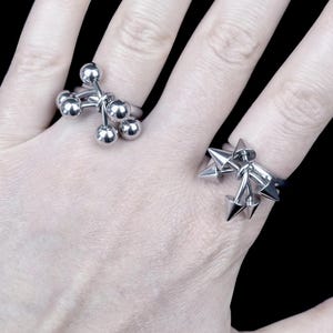 Könnte beinhalten: Zwei silberne Ringe werden auf einer Hand vor schwarzem Hintergrund gezeigt. Ein Ring hat eine Ansammlung kleiner Kugeln, der andere ein Design aus spitzen Stacheln. Die Ringe sind aus glänzendem Metall.