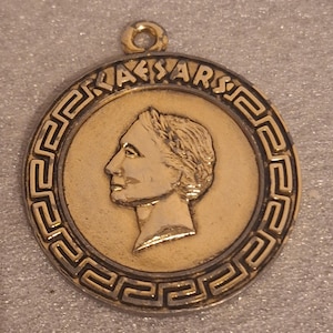 May include: A gold-coloured pendant with a profile of a person, possibly a historical figure, inside a circular frame. The word "CAESARS" is inscribed around the edge. The pendant has a loop at the top for hanging.