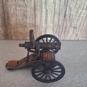 May include: A miniature bronze-colored cannon with two spoked wheels. The cannon barrel is cylindrical, and the carriage has a rectangular base. The wheels are black, and the overall design suggests a historical or antique theme.