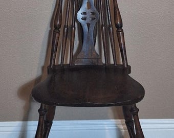 May include: Dark brown wooden chair with a spindle back and a curved top. The chair has a solid seat and four legs. The back features a decorative central element. The chair is likely antique or vintage.